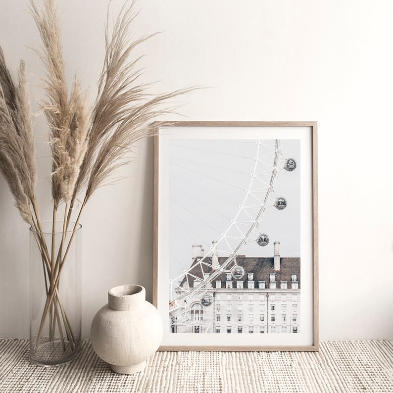 May include: A framed black and white photograph of the London Eye Ferris wheel with a building in the foreground. The photo is printed on a white background and has a light gray tint.