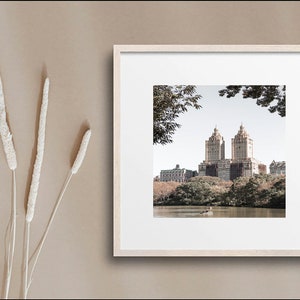 May include: A framed photograph of a city skyline with two tall buildings in the center, surrounded by trees and a lake. The photo is in a light wood frame and has a white mat.