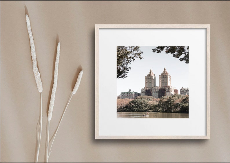 May include: A framed photograph of a city skyline with two tall buildings in the center, surrounded by trees and a lake. The photo is in a light wood frame and has a white mat.