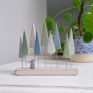 Pu&ograve; includere: Una scena di foresta in legno con alberi dai colori pastello su steli di filo metallico, un piccolo coniglio grigio e una statuina di uccello. Gli alberi sono dipinti in tonalit&agrave; di verde, blu e bianco, montati su una base di legno sbiancato. Arredamento per la casa.