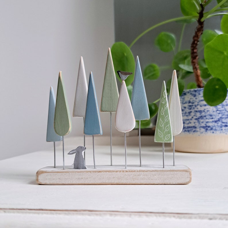 Pu&ograve; includere: Una scena di foresta in legno con alberi dai colori pastello su steli di filo metallico, un piccolo coniglio grigio e una statuina di uccello. Gli alberi sono dipinti in tonalit&agrave; di verde, blu e bianco, montati su una base di legno sbiancato. Arredamento per la casa.