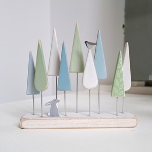 Pu&ograve; includere: Una scena di foresta in legno con alberi dai colori pastello, un piccolo coniglio grigio e un uccello. Gli alberi sono in varie tonalit&agrave; di blu, verde e bianco, montati su sottili aste metalliche e una base di legno bianca.