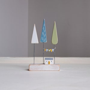 May include: A wooden sculpture of a small house with a blue roof and a yellow door, surrounded by three trees. The trees are painted white, blue with white leaves, and green. A string of colorful flags hangs between the trees.