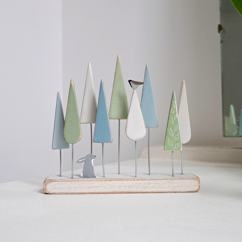 May include: A wooden forest scene with a grey rabbit, a bird, and green and blue trees. The trees are painted in various shades of green and blue and are arranged on a white wooden base.