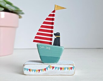 Op de afbeelding: Een houten figuur van een zwarte hond die in een turquoise zeilboot met een rood-wit gestreept zeil vaart. De zeilboot staat op een witte houten basis met een rij kleurrijke vlaggen.