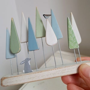 Pu&ograve; includere: Una scena di foresta in legno con alberi dipinti in tonalit&agrave; di blu, verde e bianco. Sono inclusi anche un piccolo coniglio grigio e un uccello. Gli alberi sono attaccati a una base di legno con steli di filo. Design decorativo e fantasioso.