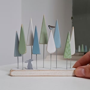 Pu&ograve; includere: Una scena di foresta in legno con vari alberi dai colori pastello su steli di filo, un coniglio grigio e un piccolo uccello. Gli alberi sono in tonalit&agrave; di blu, verde e bianco, montati su una base di legno bianca. Una mano tiene la base.