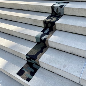 May include: A patterned rug in shades of green, black, and white is placed on a set of concrete stairs. The rug is made of a soft, textured material and has a unique abstract design.