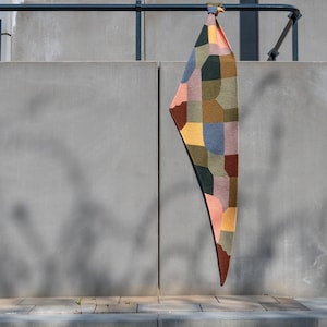 Pode incluir: Um têxtil colorido com padrão geométrico pendurado em uma barra de metal. O têxtil apresenta um design de patchwork com várias formas e cores, incluindo tons de rosa, azul, verde e amarelo. O item tem formato triangular e parece ser feito de tecido.