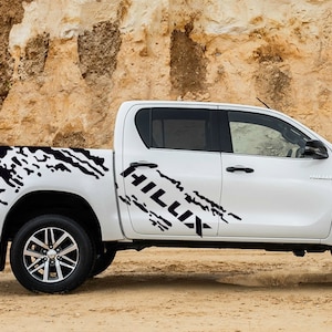 Könnte beinhalten: Weißer Pickup mit einem schwarz-weißen Grafikaufkleber an der Seite, auf dem "HILLUX" steht.