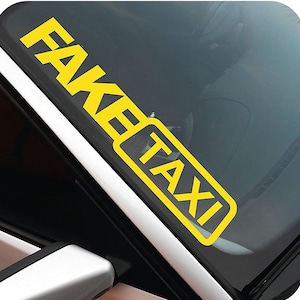 Peut inclure: Autocollant jaune "FAKE TAXI" appliqué sur le pare-brise d'une voiture. Le texte est en gras, sans empattement, avec le mot "TAXI" dans un rectangle jaune. L'autocollant est en diagonale sur la vitre.