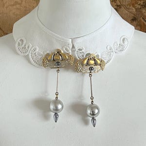 May include: A pair of gold-toned earrings featuring a basket design with fruit and a dangling gray pearl. The earrings are set against a white background, showcasing their intricate details and elegant style.