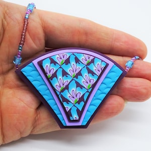 Pode incluir: Um colar com um pingente de argila polimérica com padrão roxo, azul e verde. O pingente tem a forma de um leque e um design em camadas. O colar é feito com pequenas contas.