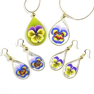May include: Jewelry set featuring teardrop-shaped pendants and earrings with colorful pansy flower designs. The pendants hang from gold-colored chains, and the earrings have gold-colored hooks. The pansies have yellow, purple, blue, and red petals.