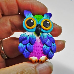 May include: A colorful owl figurine made of polymer clay. The owl has blue, purple, pink, green, and orange accents. The owl's eyes are large and black with orange rings around them.