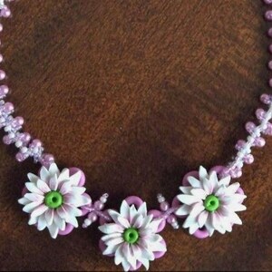 May include: A delicate necklace made with white and light purple beads, featuring three white flower charms with green centres.