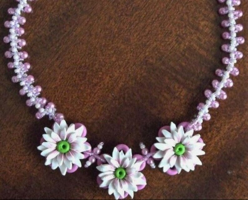 May include: A delicate necklace made with white and light purple beads, featuring three white flower charms with green centres.