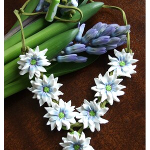 May include: A white flower necklace with green centres and blue accents. The necklace is made of polymer clay and is strung on a brown suede cord. The necklace is lying on a wooden surface with a bouquet of purple hyacinths.