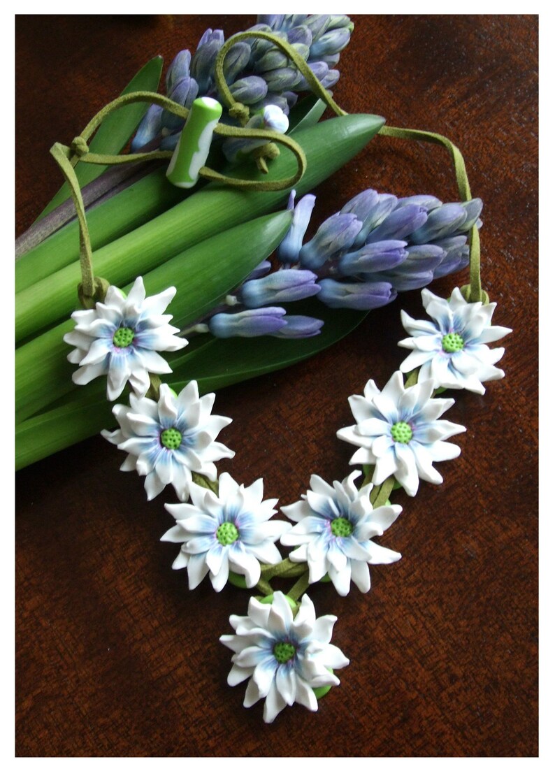 May include: A white flower necklace with green centres and blue accents. The necklace is made of polymer clay and is strung on a brown suede cord. The necklace is lying on a wooden surface with a bouquet of purple hyacinths.