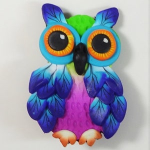 May include: A colorful owl figurine made of polymer clay. The owl has bright blue wings, a green head, and large orange eyes with black pupils. The owl's body is pink and has a textured surface.