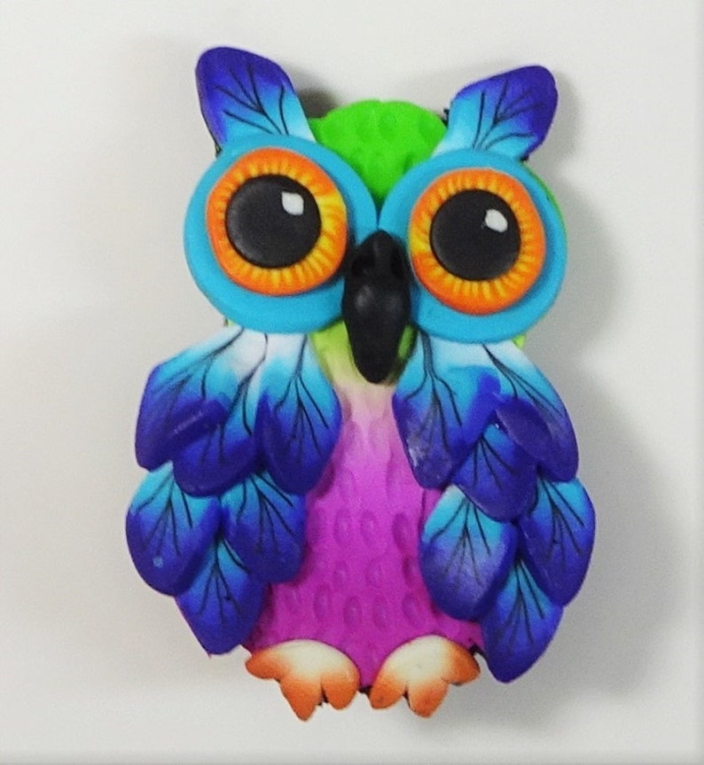 May include: A colorful owl figurine made of polymer clay. The owl has bright blue wings, a green head, and large orange eyes with black pupils. The owl's body is pink and has a textured surface.