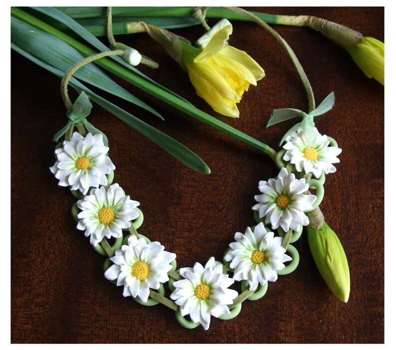 May include: A white daisy flower necklace with green leaves and white beads. The necklace is made of polymer clay and is a handmade piece of jewellery.
