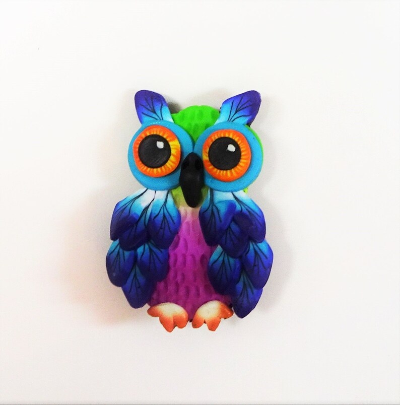 May include: A colorful owl figurine made of polymer clay. The owl has bright blue and purple wings, green and orange eyes, and a pink body.