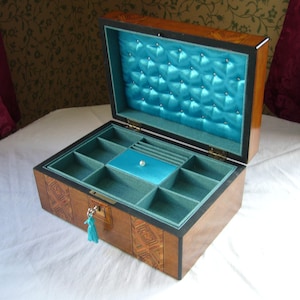 May include: A rectangular wooden jewelry box with a polished finish and geometric inlay patterns. The interior features a tufted bright turquoise satin lid lining and a teal velvet-lined base with multiple compartments and a central lidded section.