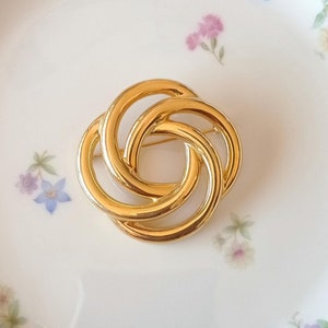 May include: A gold-toned metal brooch in the shape of a knot. The brooch is made of three interlocking circles.