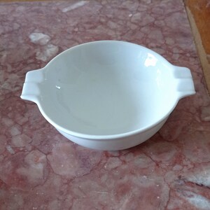 May include: A white ceramic bowl with two handles. The bowl is empty and sitting on a pink and white marble surface.