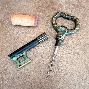 Può includere: Un cavatappi vintage a forma di chiave con finitura verde e oro. Il cavatappi ha un manico decorativo con un motivo floreale e una vite in metallo argentato.