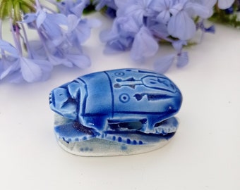 Puede incluir: Una figurita de escarabajo de cerámica azul con detalles intrincados. El escarabajo está sentado sobre una superficie blanca.