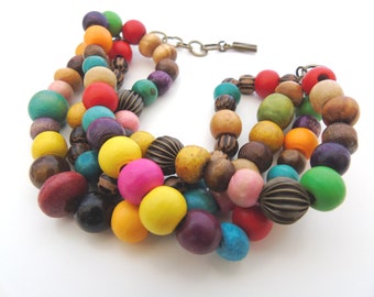 Puede incluir: Un collar de múltiples hilos hecho de cuentas de madera de colores. Las cuentas son de varias formas y tamaños, incluyendo redondas, ovaladas y cilíndricas. El collar es una mezcla vibrante de colores, incluyendo rojo, naranja, amarillo, verde, azul, morado y marrón.