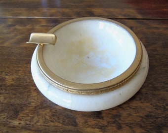 May include: A vintage ashtray with a white interior and a gold-colored metal rim and cigarette rest. The ashtray has a rounded shape and sits on a wooden surface. The interior shows signs of use.