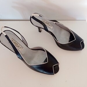 May include: A pair of black leather heeled sandals with a white trim. The sandals have a slingback strap and an open toe. The brand name "GRECAS" is printed on the insole.