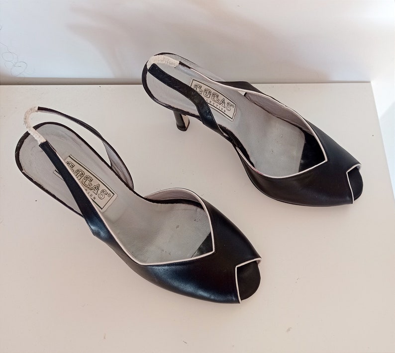 May include: A pair of black leather heeled sandals with a white trim. The sandals have a slingback strap and an open toe. The brand name "GRECAS" is printed on the insole.