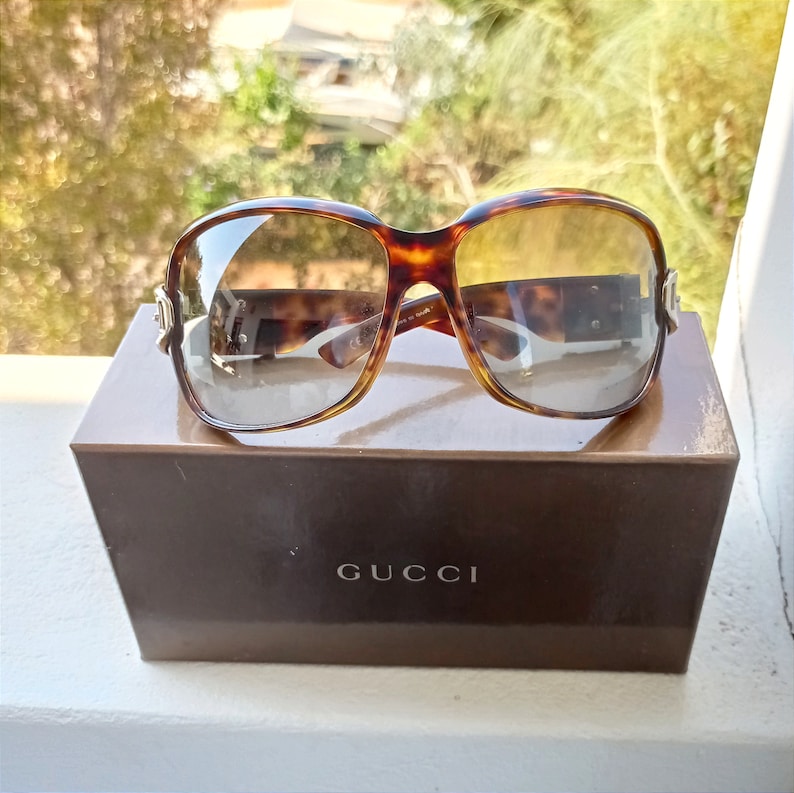 Puede incluir: Gafas de sol de carey marr&oacute;n con un acento plateado en el lateral. Las gafas de sol est&aacute;n sobre una caja marr&oacute;n con la palabra "GUCCI" impresa en la parte delantera.