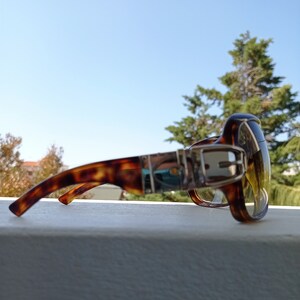 Puede incluir: Gafas de sol de carey con un detalle de hebilla plateada en el marco. Las lentes son de color amarillo.