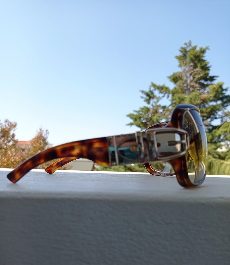 Puede incluir: Gafas de sol de carey con un detalle de hebilla plateada en el marco. Las lentes son de color amarillo.