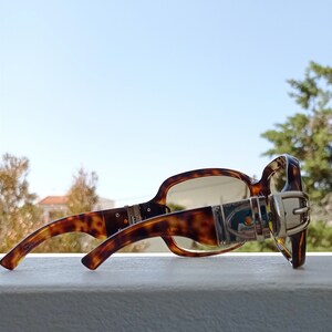 Puede incluir: Gafas de sol de carey con un detalle de hebilla plateada en el lateral. Las gafas tienen lentes marrones y est&aacute;n apoyadas sobre una superficie blanca.