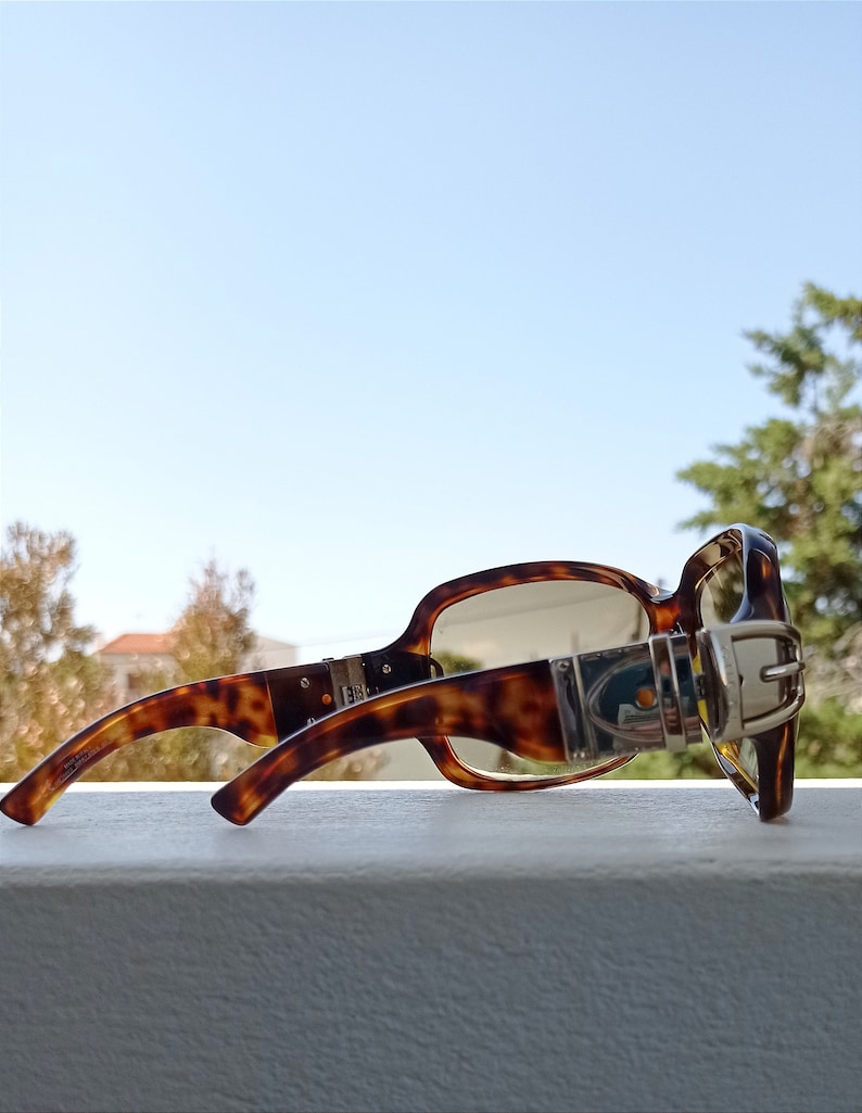 Puede incluir: Gafas de sol de carey con un detalle de hebilla plateada en el lateral. Las gafas tienen lentes marrones y est&aacute;n apoyadas sobre una superficie blanca.
