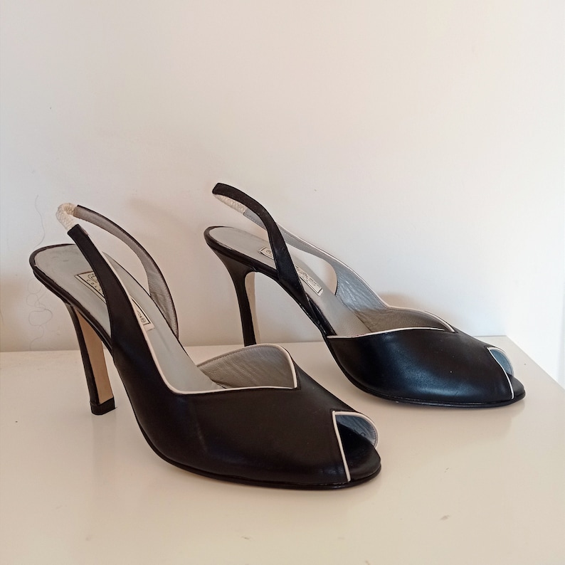 May include: A pair of black leather slingback heels with a white trim. The heels are open-toed and have a high stiletto heel.
