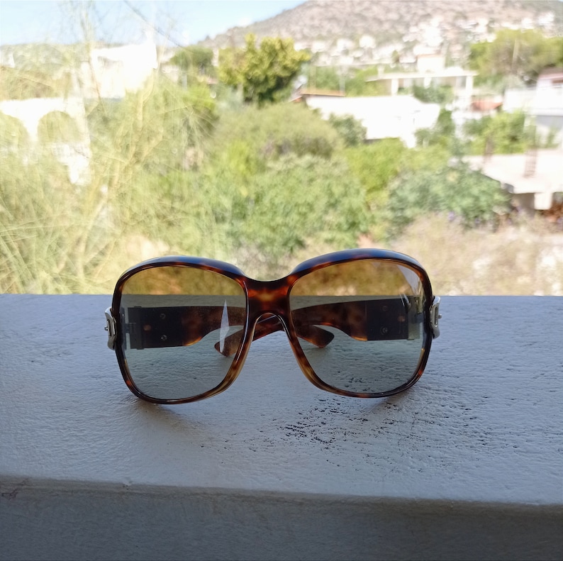 Puede incluir: Un par de gafas de sol de carey marr&oacute;n con un acento plateado en el lateral. Las gafas de sol est&aacute;n sobre una superficie blanca.