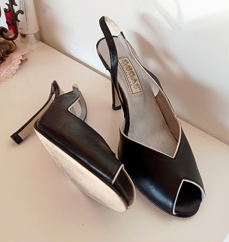 May include: A pair of black peep-toe heels with a white trim. The heels are made of leather and have a slingback strap. The shoes are labeled "GORE-TEX COLLECTION".