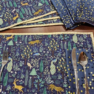 May include: A set of navy blue placemats and napkins with a whimsical forest design. The placemats feature deer, leopards, and peacocks among trees and foliage. Napkins have a navy blue background with yellow polka dots. A fork, knife, and spoon are visible.
