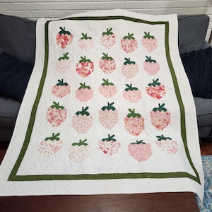 May include: A white quilt featuring a pattern of pink strawberries with green stems and leaves. The strawberries have various floral patterns. The quilt has a green border and is displayed on a dark brown table.