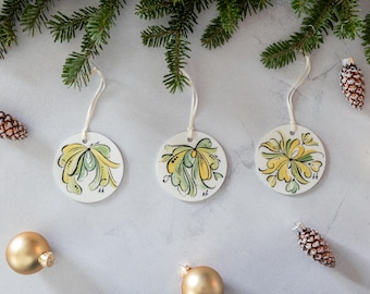 Op de afbeelding: Drie ronde, witte keramische ornamenten met gele en groene bloemenmotieven. Elk ornament heeft een touwtje om op te hangen.