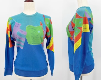 Puede incluir: Un suéter azul de manga larga con un diseño abstracto colorido con banderas y estrellas. El suéter tiene un cuello y puños de canalé.