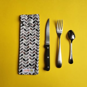 May include: A cutlery set arranged on a yellow surface. The set includes a knife, fork, and spoon, alongside a fabric utensil holder. The holder is white with a pattern of black, brown, and grey leaves. The knife has a black handle.