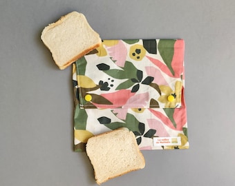 May include: A reusable sandwich wrap with a floral pattern in pink, green, yellow, and black. Two slices of bread are next to the wrap. The wrap has yellow snap closures and a small label with text.